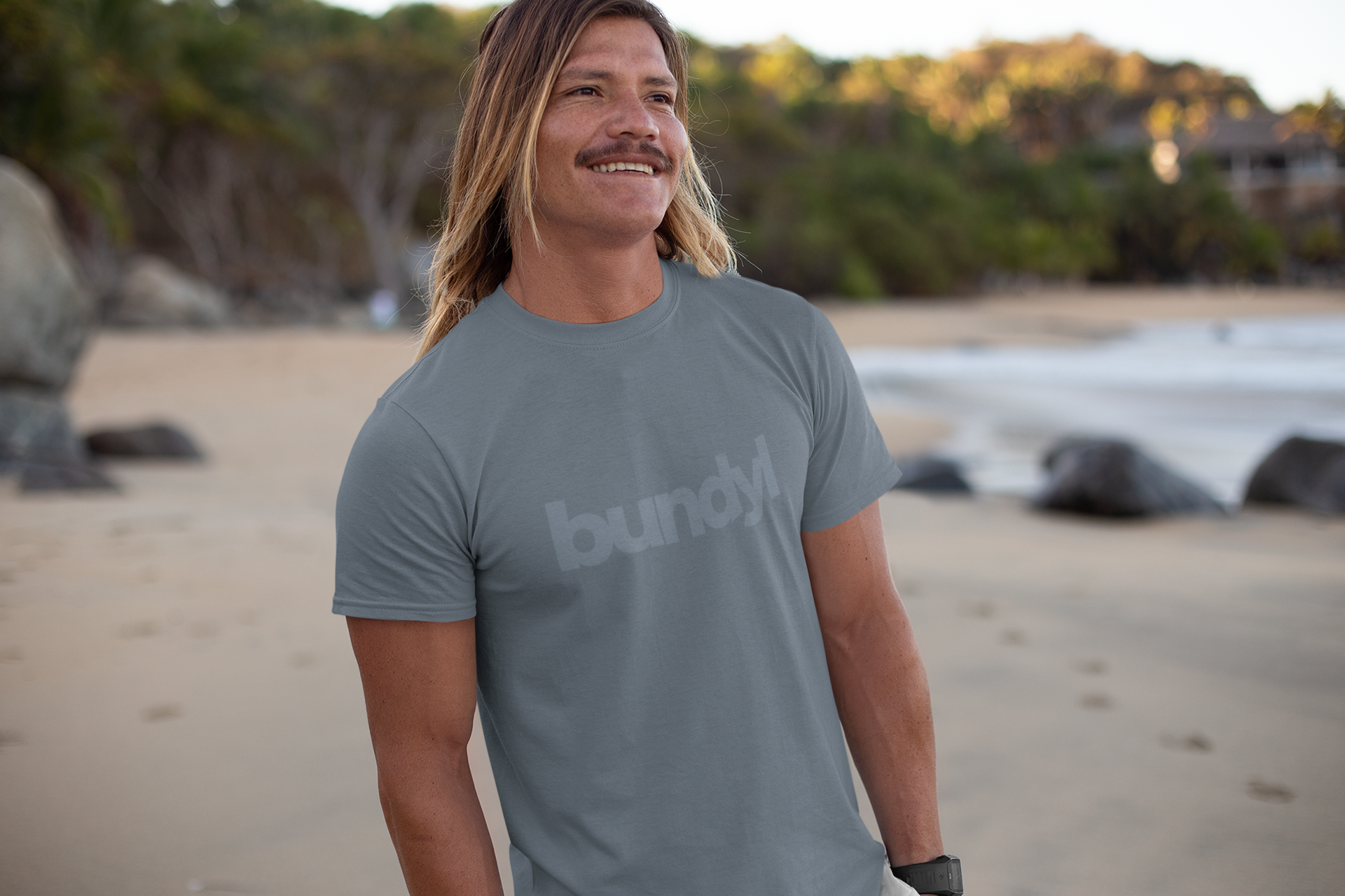 Man wearing a gray 'bundy' t-shirt on a beach with trees and water in the background